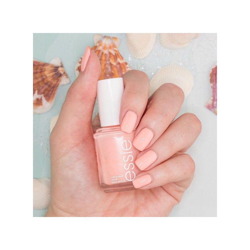 Hand met gelakte nagels en nagellakfles. Fles met witte dop, roze lak. Op lichte achtergrond met schelpen.