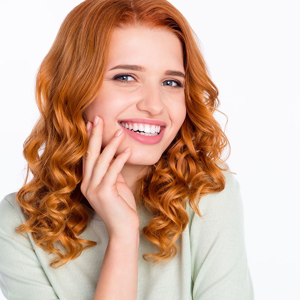 Portret van een lachende vrouw met rood krullend haar, die haar wang aanraakt.