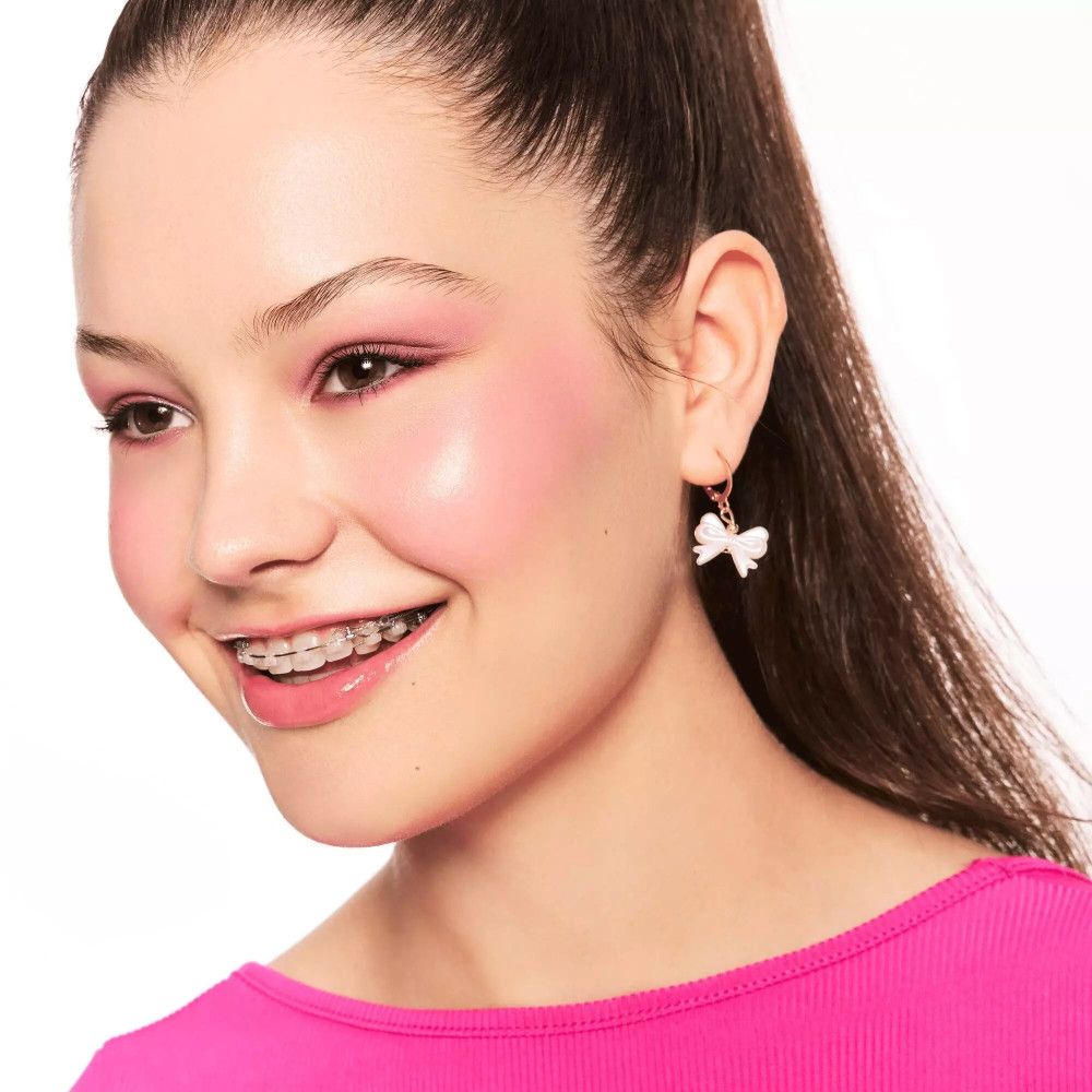 Portret van een lachende vrouw met roze wangen. Ze draagt een roze blouse en strikvormige oorbellen.