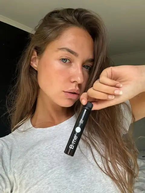 Vrouw houdt een zwarte ampul vast met een witte stip en B.me®-logo. Ze draagt een grijs T-shirt.