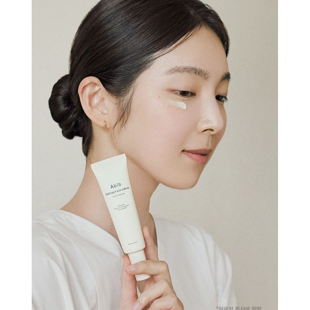 Vrouw houdt een lichtbeige crèmetube vast. Opschrift: Abib Collagen eye crème. Ze brengt de crème aan onder het oog.