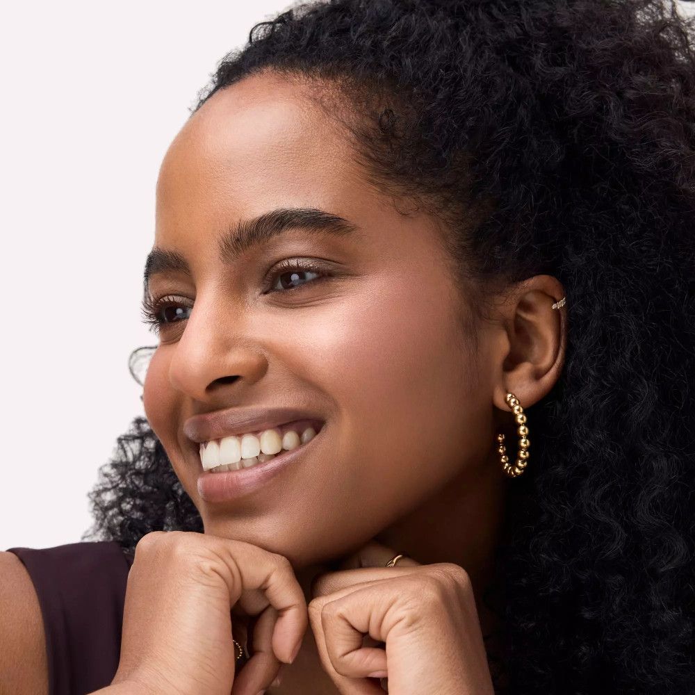 Portrait d'une femme souriante avec des cheveux foncés et des boucles d'oreilles dorées. Elle soutient son menton avec ses mains.