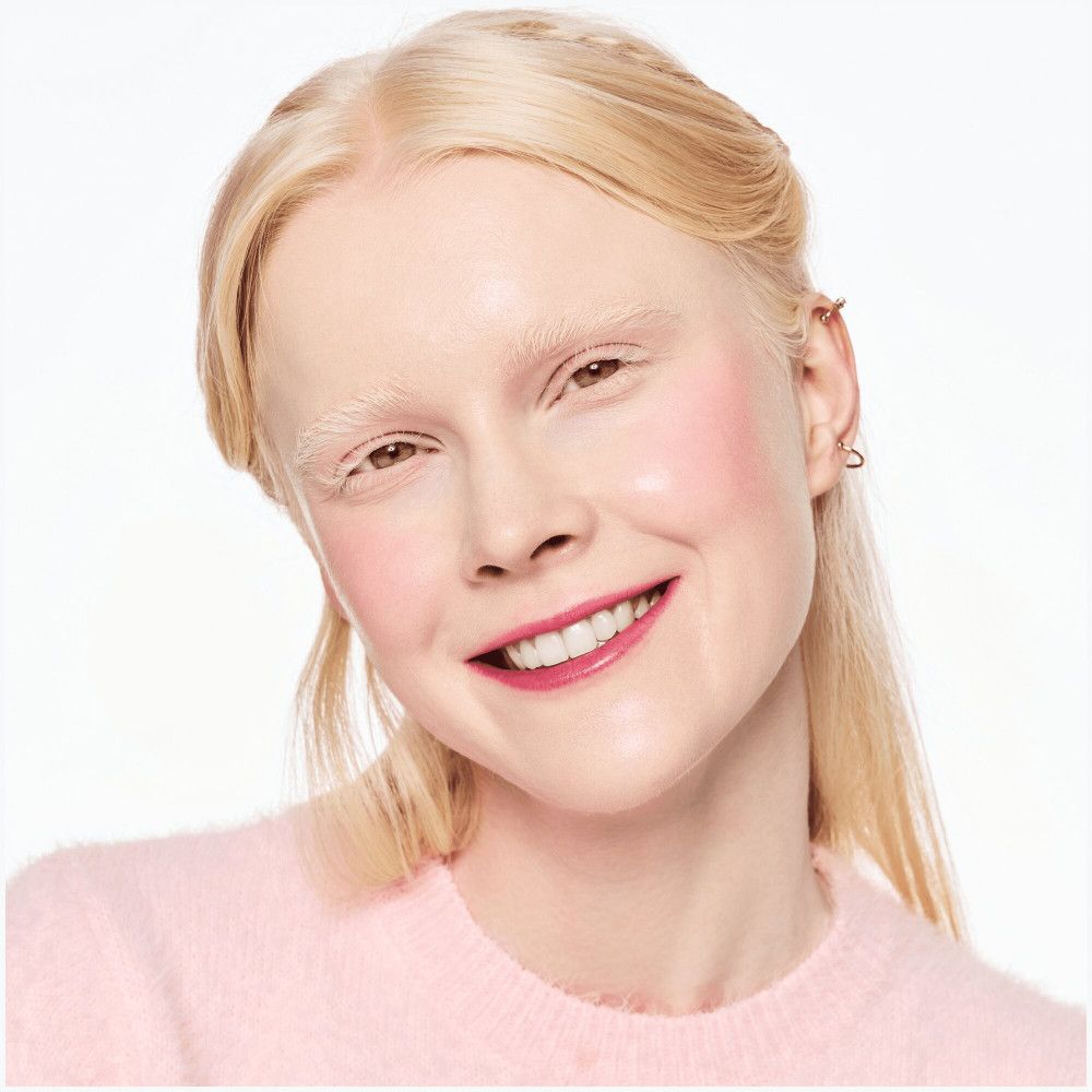 Blonde vrouw met roze getinte wangen en lippen. Ze draagt een roze trui en glimlacht.