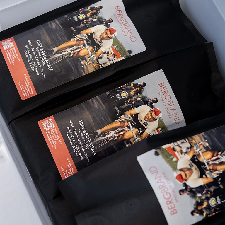 Drie zwarte koffiebonenzakken met etiket. Opdruk: BERGBRAND, Eddy Would Attack, fietser illustratie.