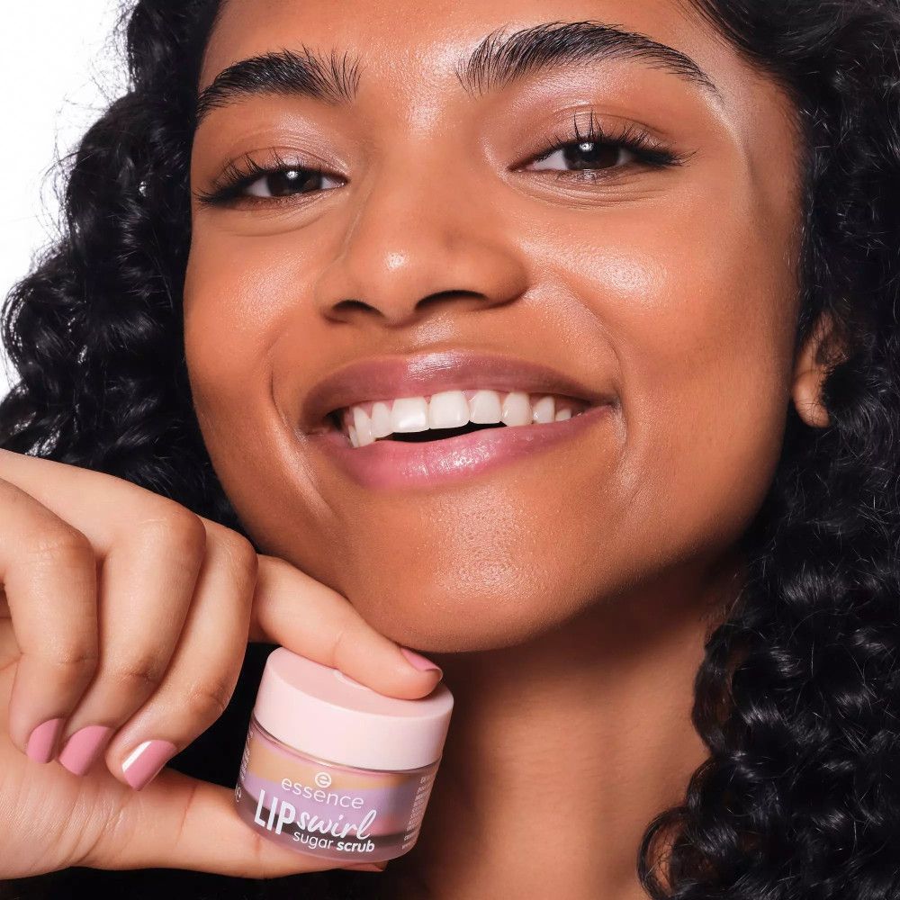 Personne tenant un gommage pour les lèvres. Couvercle rose avec logo blanc. Nom du produit visible. La personne sourit.