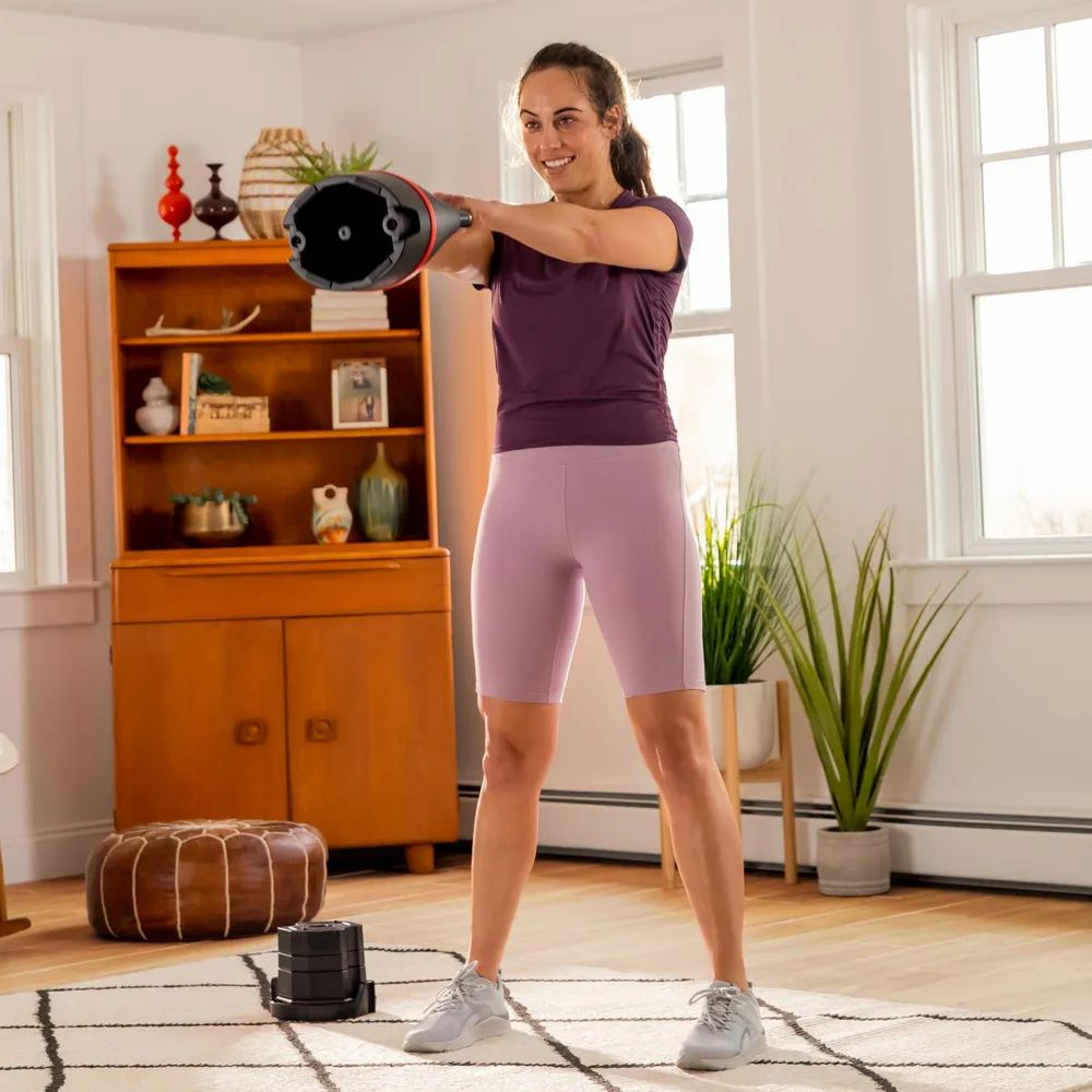 Femme avec kettlebell. Haltère russe noire avec bande rouge. Logo Bowflex visible.