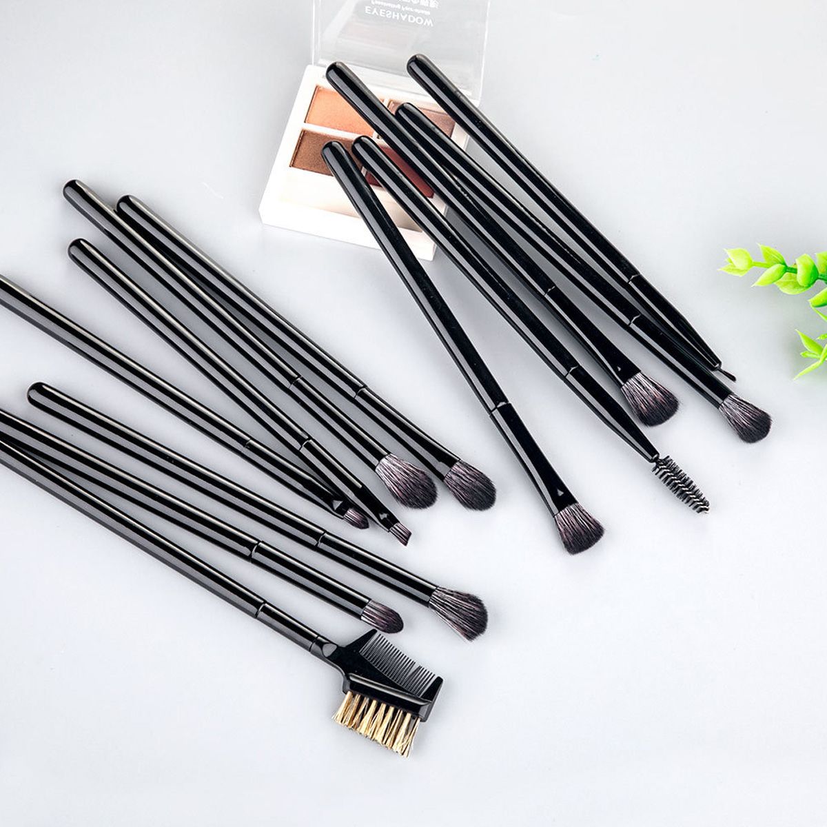 Pinceaux noirs pour fard à paupières et sourcils, partiellement sur une surface grise, avec un set de fard à paupières en arrière-plan.