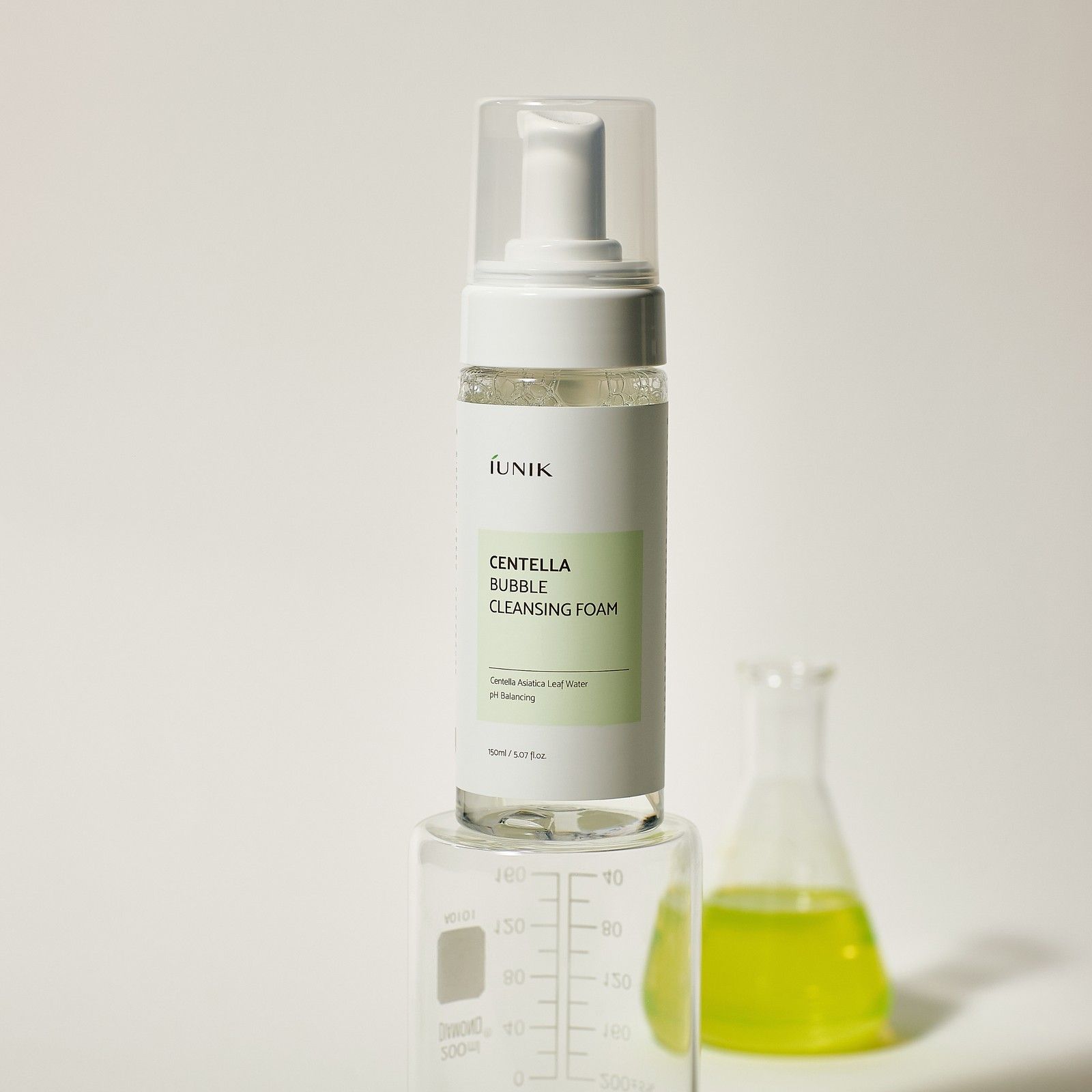 iUNIK Centella Bubble Cleansing Foam fles op een maatcilinder. Een erlenmeyer met gele vloeistof staat ernaast.