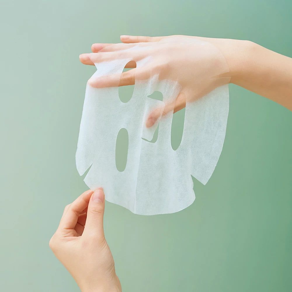 Mains tenant un masque facial blanc. Le masque est transparent et de forme ovale. Le fond est vert.