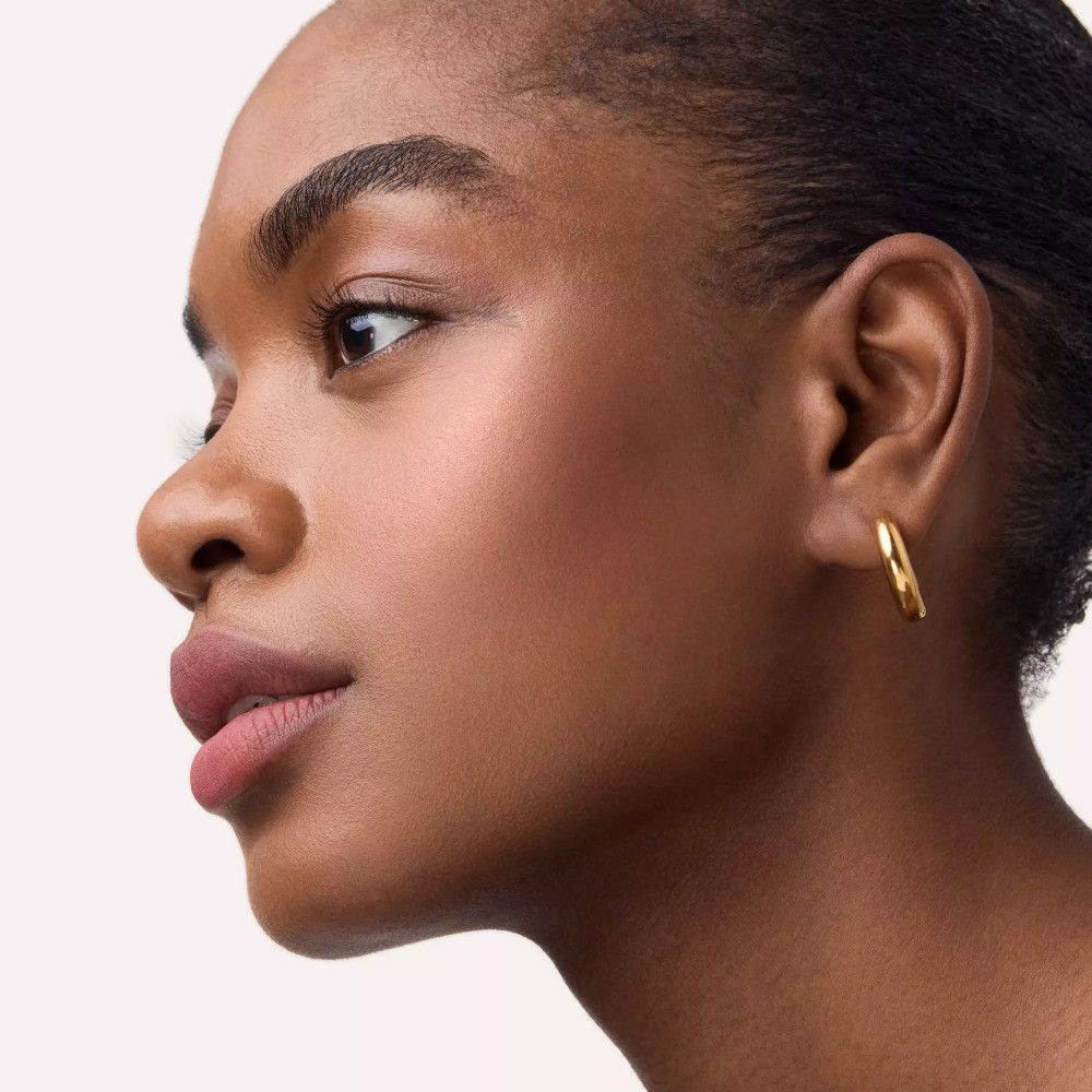 Close-up van een vrouw met een gebruinde huid en blush op de wangen. Ze draagt gouden oorringen.