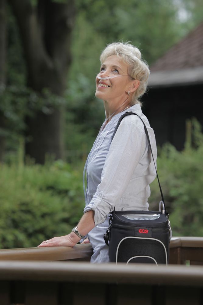Vrouw met GCE Zen-O zuurstofconcentrator. Zwarte tas met schouderriem. Neusbril.