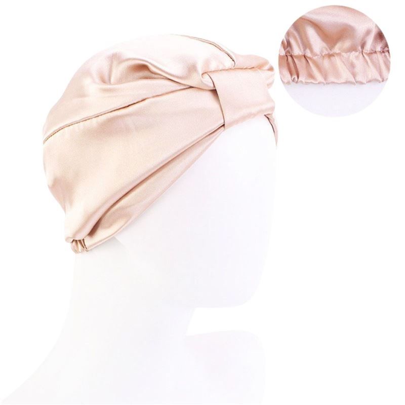 Un bonnet de nuit en soie de couleur rose pâle. Le bonnet a une forme de turban avec un nœud décoratif à l'avant. Le matériau semble lisse et brillant. Un petit encart montre le bord froncé.