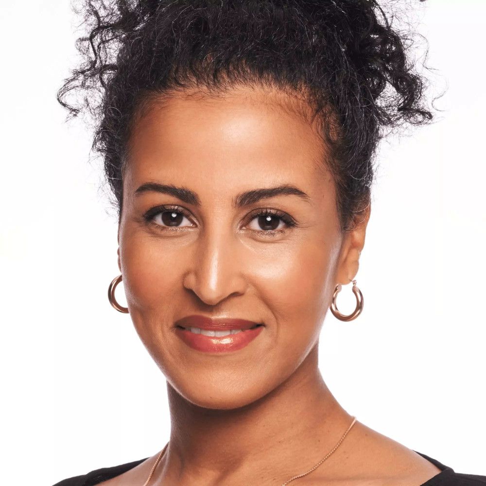 Portrait d'une femme à la peau foncée et aux cheveux bouclés. Elle porte des boucles d'oreilles dorées et un haut noir.