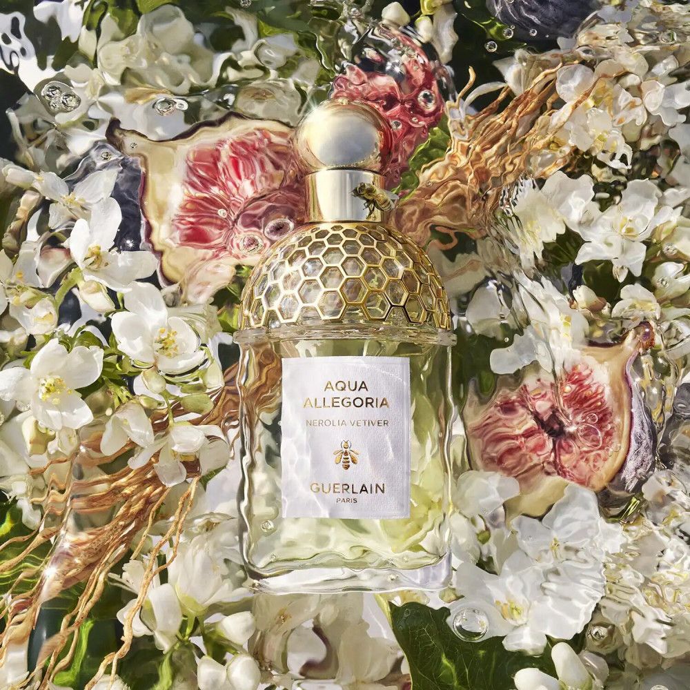 Flacon de parfum avec bouchon doré, entouré de fleurs et de fruits. Étiquette avec texte : Aqua Allegoria Nerolia Vetiver, Guerlain.