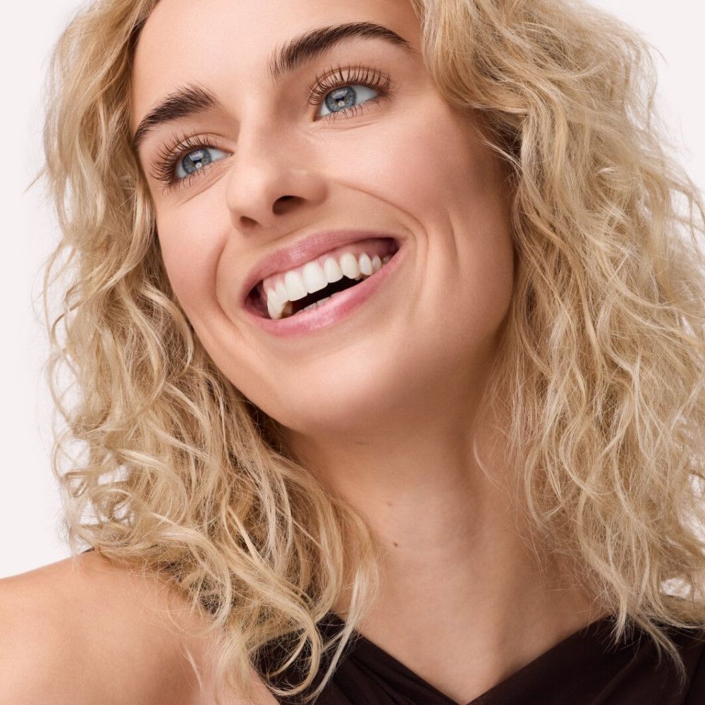 Portret van een lachende vrouw met blonde krullen en blauwe ogen. Ze draagt donkere kleding.