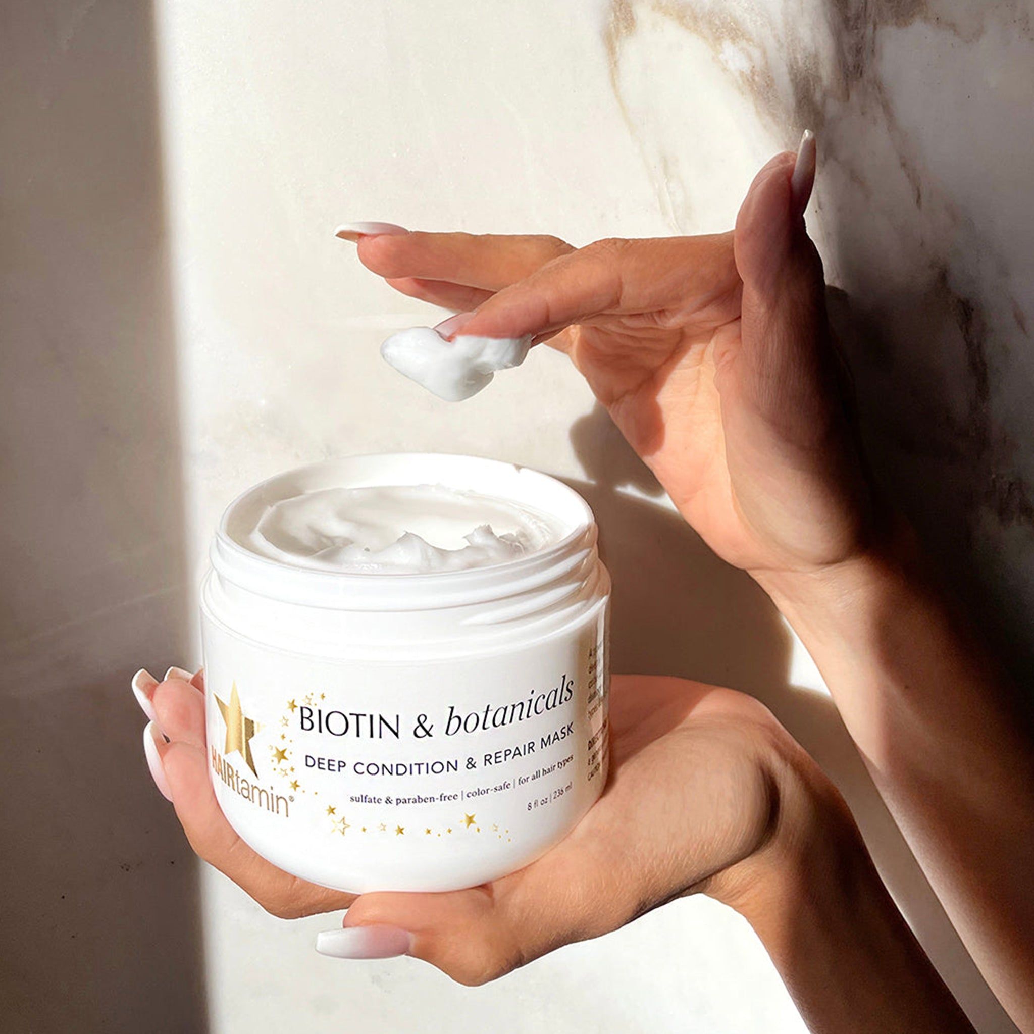 Mains tenant un pot de masque capillaire et prélevant du produit. Texte: BIOTIN & botanicals DEEP CONDITION & REPAIR MASK.
