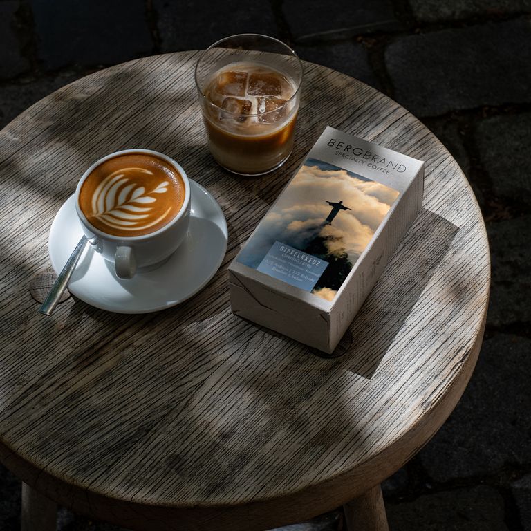 Café et boisson sur table. Paquet BERGBRAND Gipfelkreuz. Tasse à café avec latte art. Verre avec glaçons.