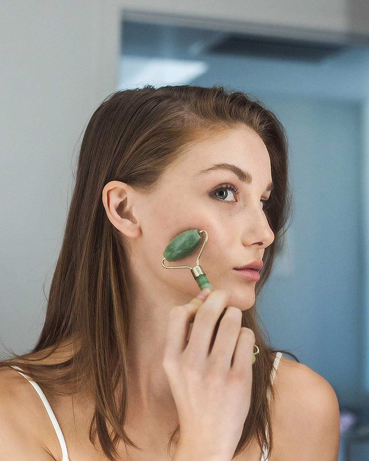 Une femme aux cheveux bruns utilise un rouleau facial en jade vert sur sa joue.