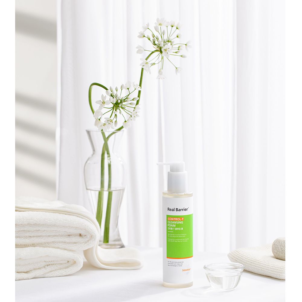Wit-groene fles met pomp naast bloemen en handdoeken. Tekst: Real Barrier, Control-T Cleansing Foam. Kom met vloeistof.