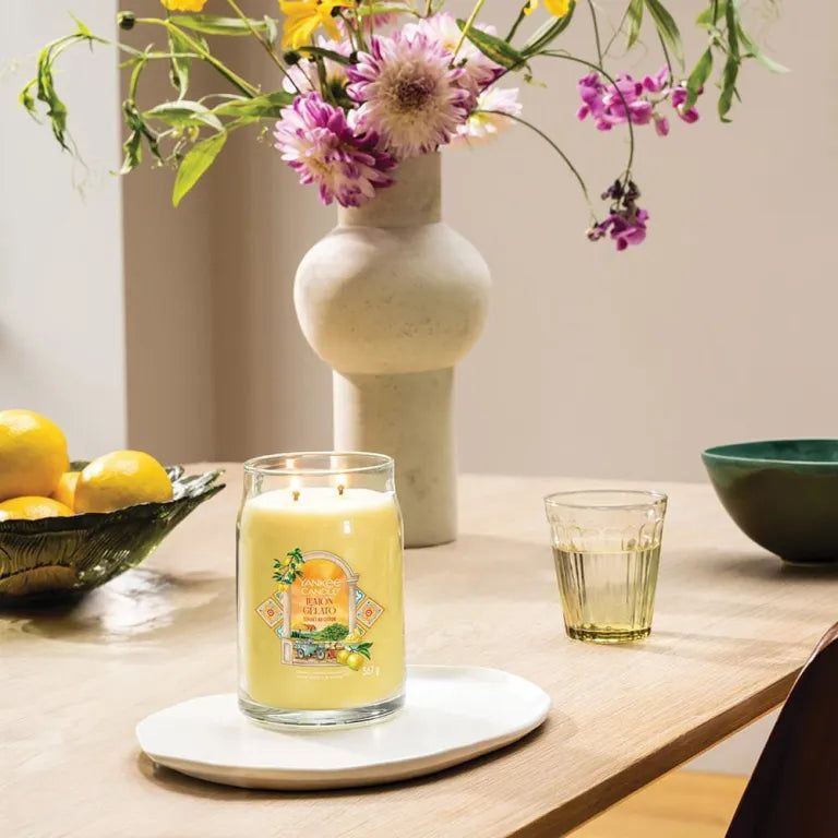 Grand pot de bougie jaune avec deux mèches et étiquette. Sur une assiette blanche. Fond : table, fleurs, verre.