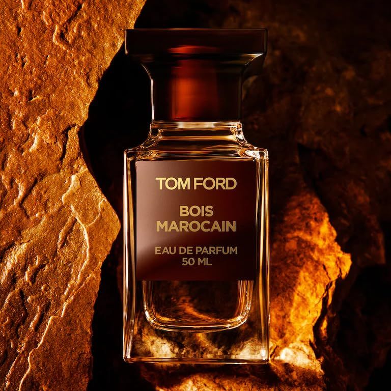 Glazen fles met bruine dop. Opschrift: TOM FORD, BOIS MAROCAIN, EAU DE PARFUM, 50 ML. Achtergrond: goudkleurige steen.