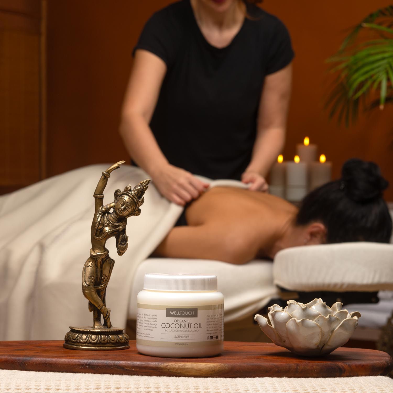 Wit glazen pot met COCONUT OIL. Massage in uitvoering. Bronzen beeld en lotusbloem op houten plank.