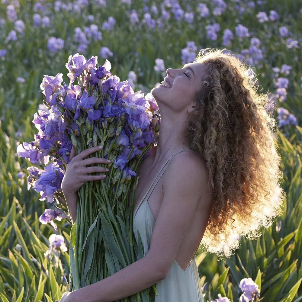 Een vrouw houdt een boeket paarse bloemen vast in een veld. Ze lacht en kijkt omhoog.