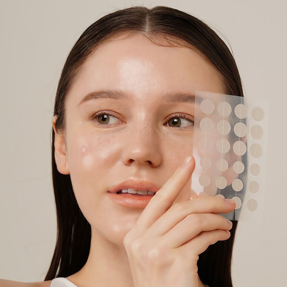 Persoon die een transparante folie met ronde pleisters vasthoudt. Een puistje is zichtbaar op het gezicht.