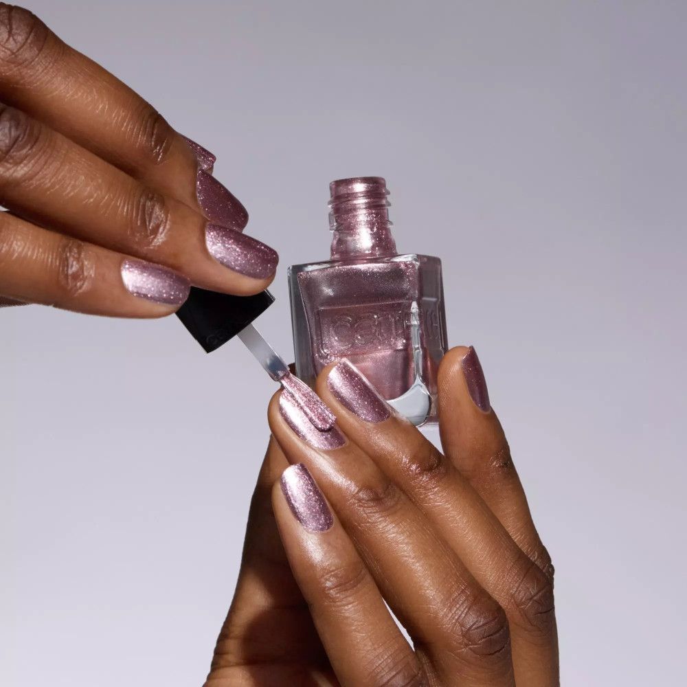 Mains tenant un flacon de vernis à ongles et un pinceau. Vernis à ongles rose scintillant sur les ongles. Flacon avec inscription "catrice".