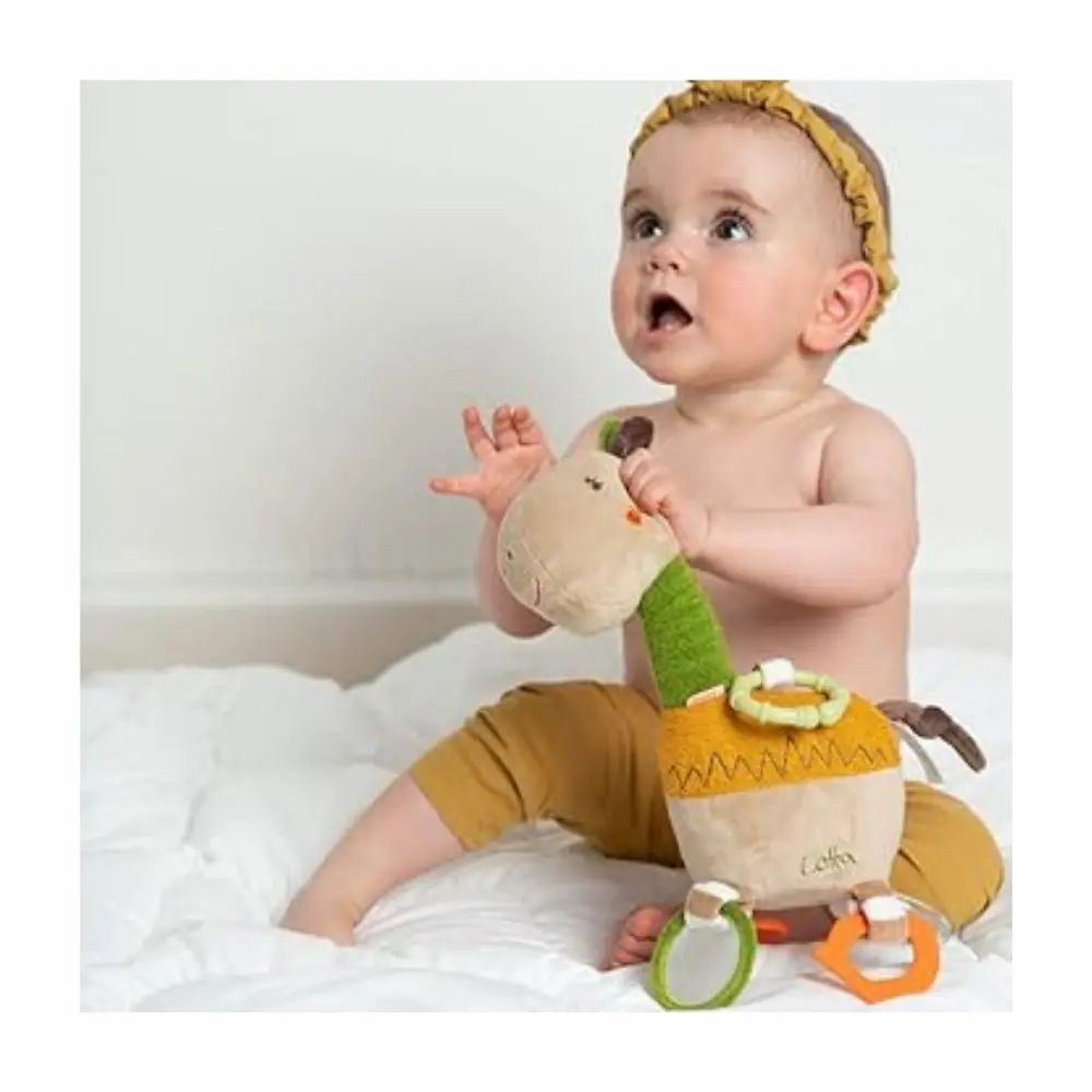 Bébé tenant une girafe en peluche. Beige, vert et jaune. Anneaux et miroirs. Inscription "Lotta". Bébé porte un bandeau.