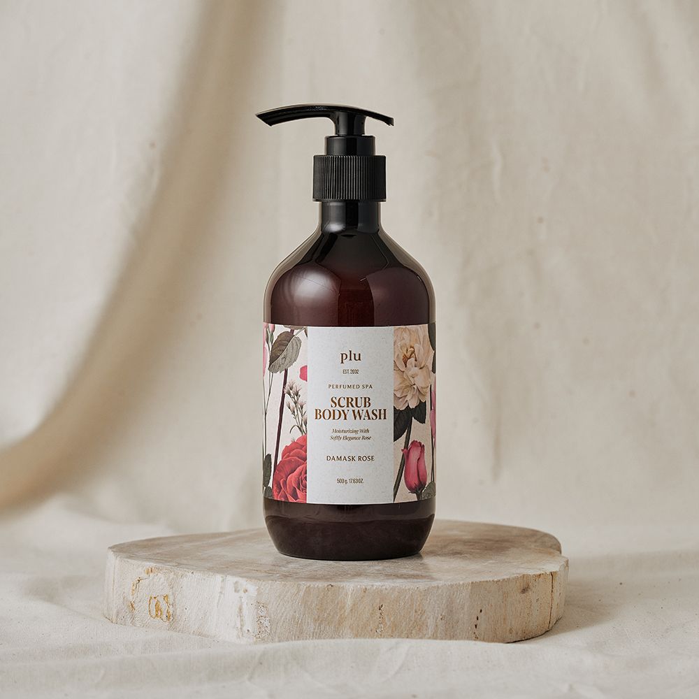 Flacon marron avec pompe noire. Étiquette florale avec texte "SCRUB BODY WASH DAMASK ROSE". Posé sur un socle.