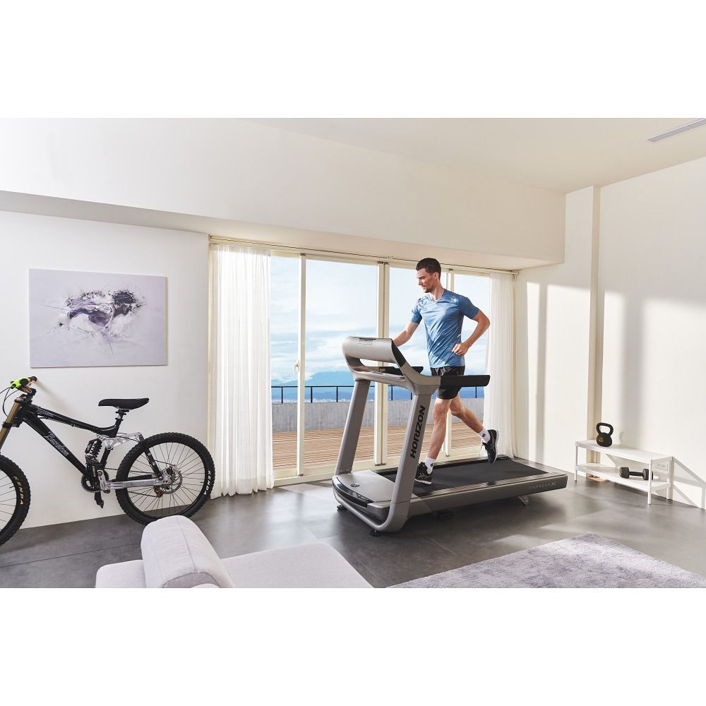 Tapis de course gris dans un salon. Le coureur s'entraîne. Visible : fenêtre, vélo, haltère, canapé. Logo Horizon sur le tapis.