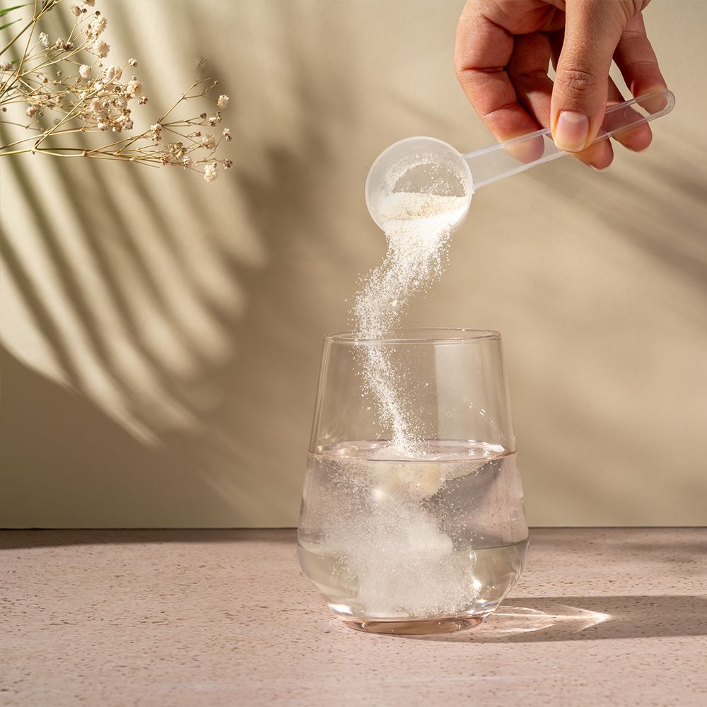 Hand schept poeder uit een maatschepje in een glas water. Achtergrond: licht, met schaduwen.