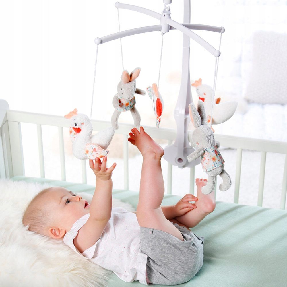 Bébé sous mobile, tendant les bras. Mobile avec lapins, cygnes et miroir. Structure blanche. Accents roses et bleus.