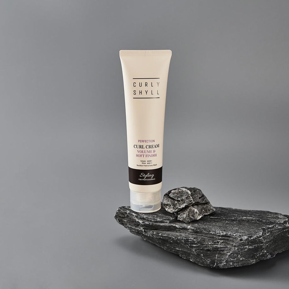 Crèmetube op een steen. Tekst: CURLY SHYLL, Perfection Curl Cream, Volume & Soft Finish. Zwarte strook met tekst Styling.
