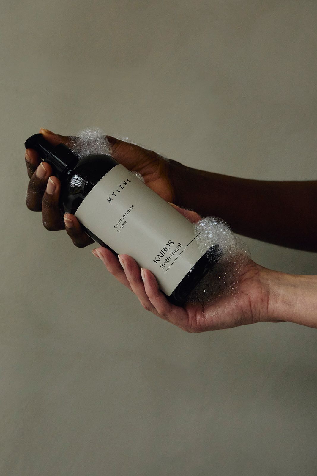 Handen houden een pompfles met licht etiket "KAIROS [Bath foam]" vast, omringd door schuim.