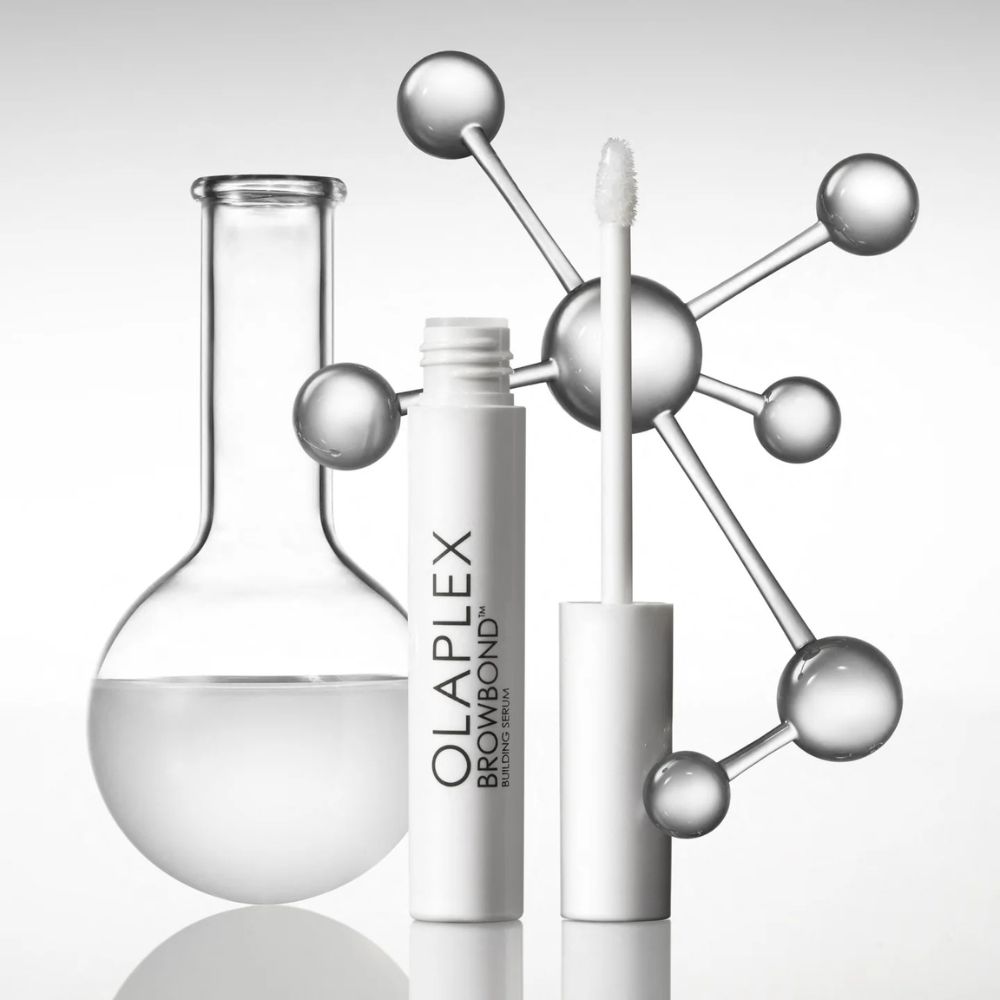 Wit serumflesje met applicator. OLAPLEX BROWBOND BUILDING SERUM. Naast een reageerbuis en een moleculaire structuur.