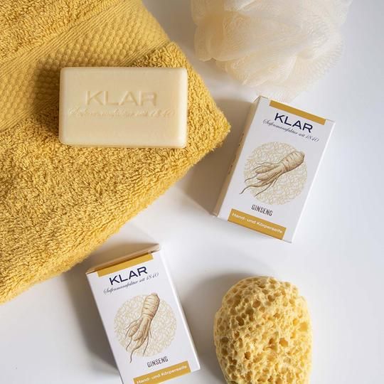 Zeep en verpakkingen op gele handdoek en sponzen. Zeep met KLAR reliëf. Verpakkingen: KLAR, Ginseng, hand- en lichaamszeep.