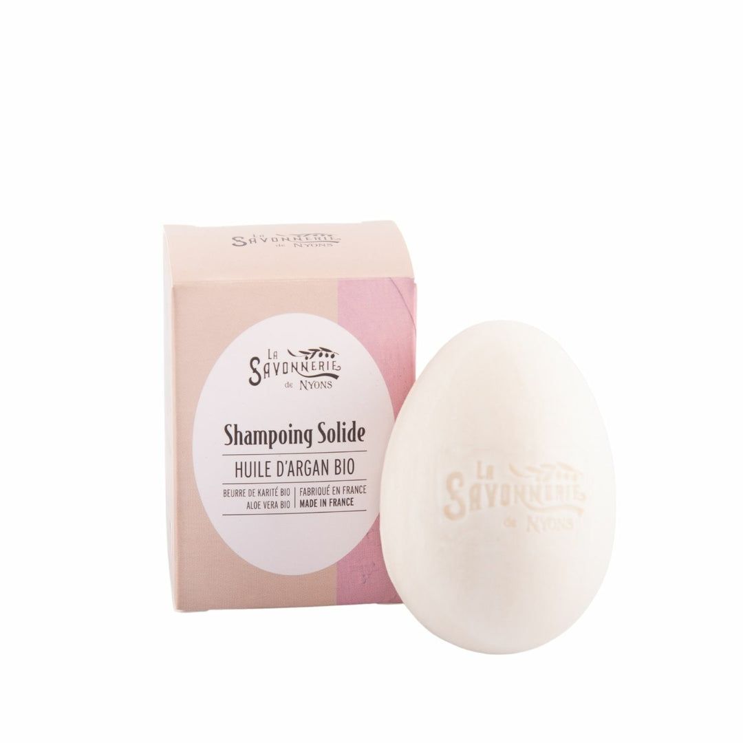 Eivormige, vaste shampoo naast kartonnen verpakking. Roze-beige kleurovergang. Wit etiket met tekst: Huile d'Argan Bio, Made in France.