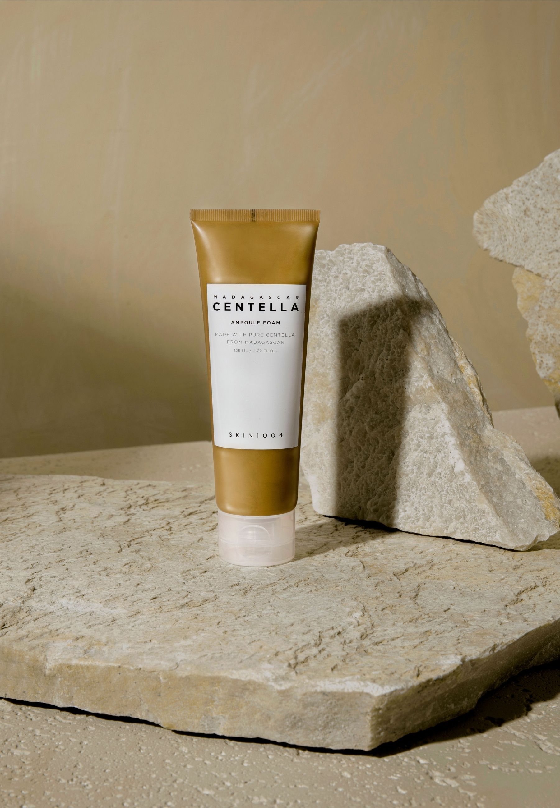 Beige tube met wit etiket. Opschrift: MADAGASCAR CENTELLA, Ampoule Foam, SKIN1004. Geplaatst op stenen.