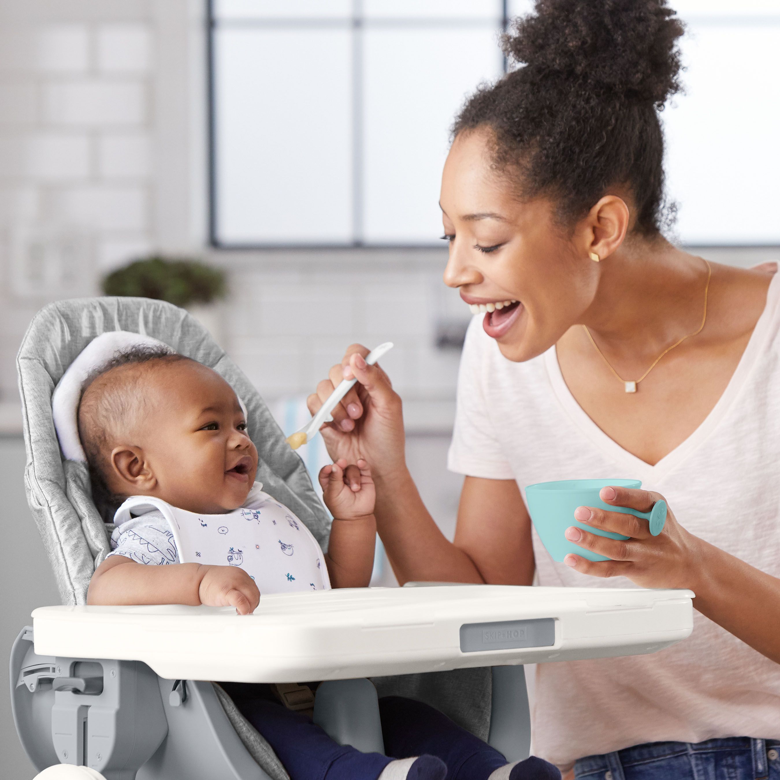 Een vrouw voedt een baby in een kinderstoel met een babyvoedingskom. De baby lacht.