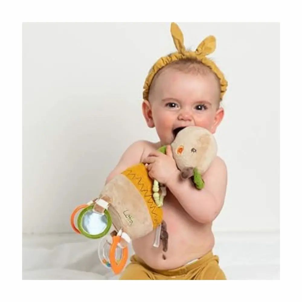 Bébé mord une girafe en peluche. Beige, vert et jaune. Anneaux et miroirs. Inscription "Lotta". Bébé porte un bandeau.