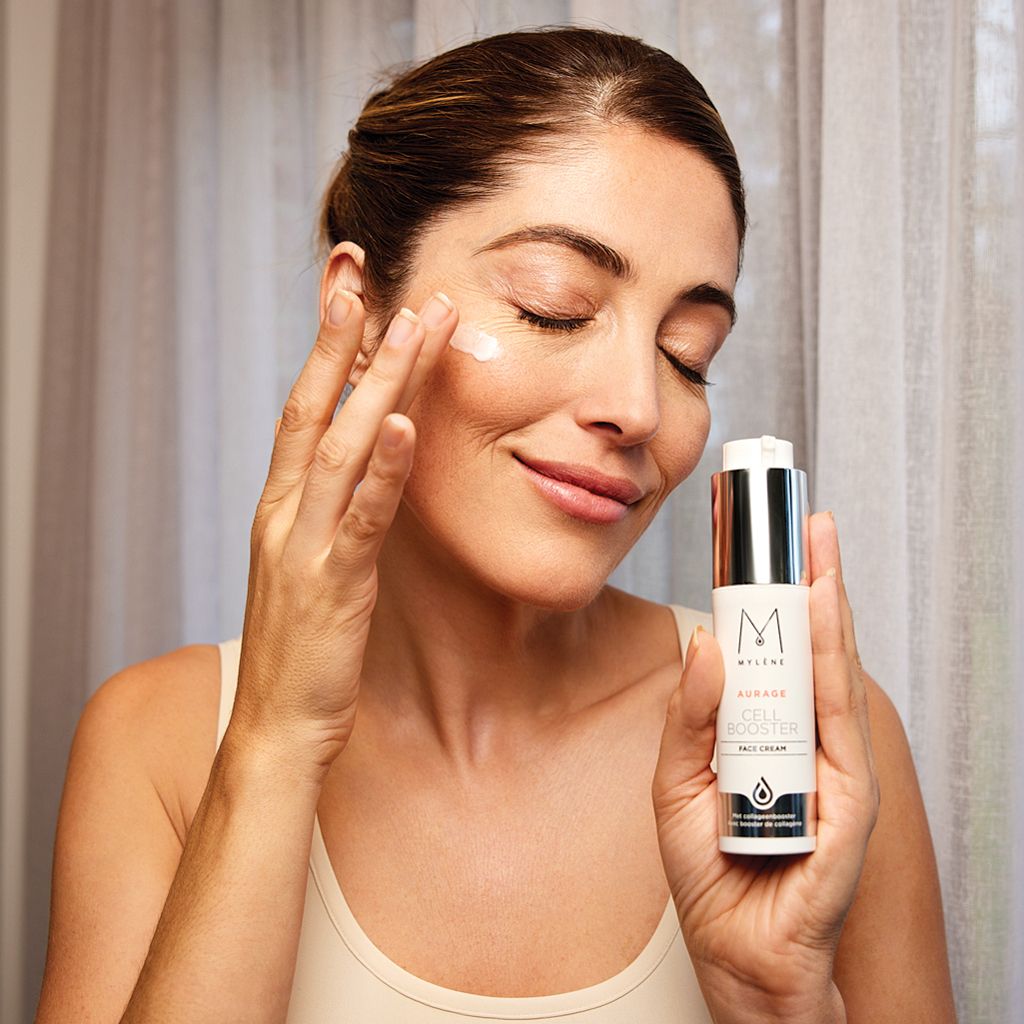 Femme appliquant de la crème. Flacon avec logo MYLÈNE et texte Aurage Cell Booster. Emballage argenté et blanc.