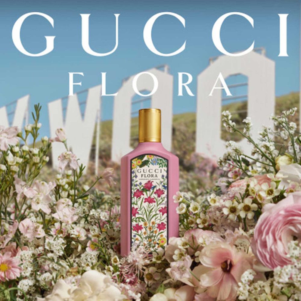 Flacon rose Gucci Flora avec bouchon doré, entouré de fleurs. En arrière-plan: panneau Hollywood. Texte: Gucci Flora.