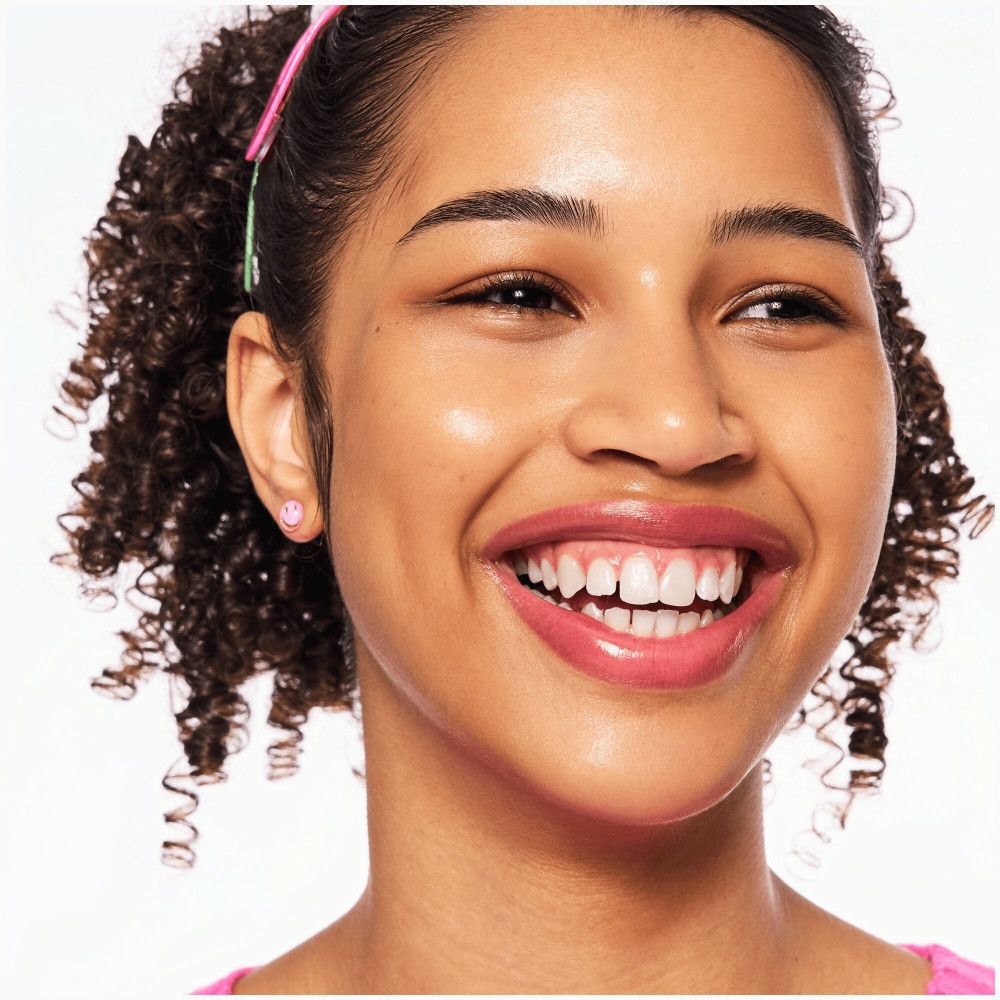 Gros plan d'un visage souriant. Rouge à lèvres rose. Cheveux bouclés. Boucle d'oreille rose. Fond clair.