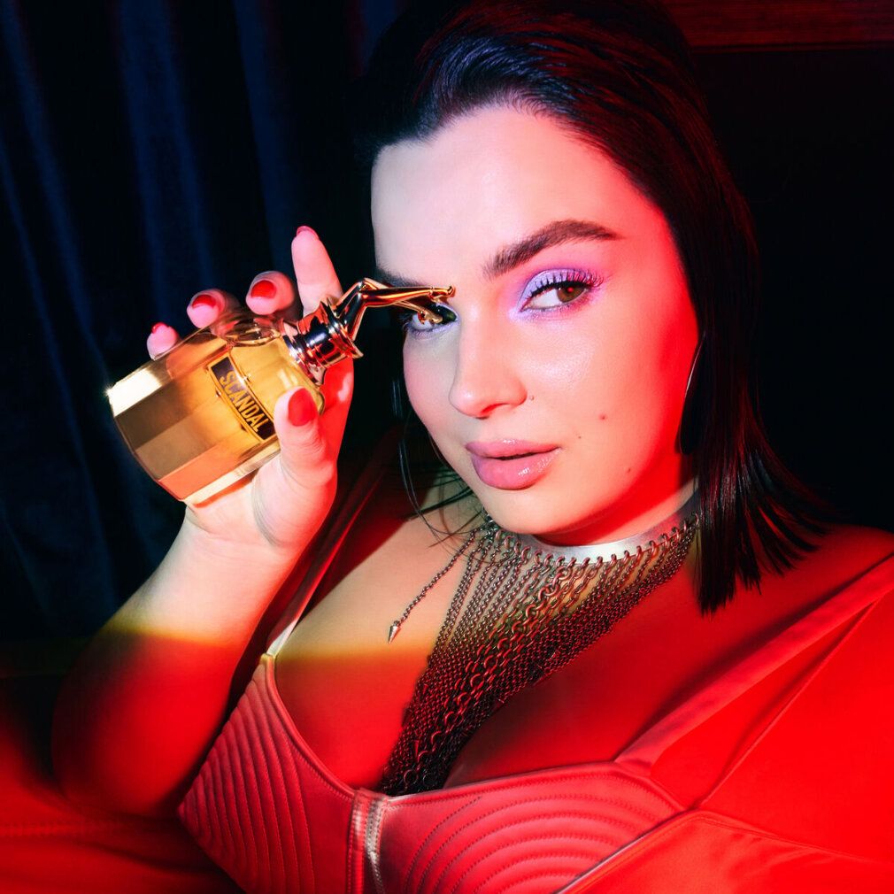 Femme tenant un flacon de parfum avec bouchon doré. Flacon avec inscription Scandal. La femme porte un haut rouge et des bijoux.