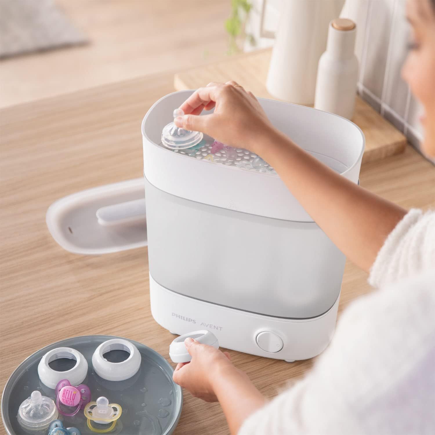 Une main place un biberon dans un stérilisateur Philips Avent, d'autres pièces sont sur un plateau.