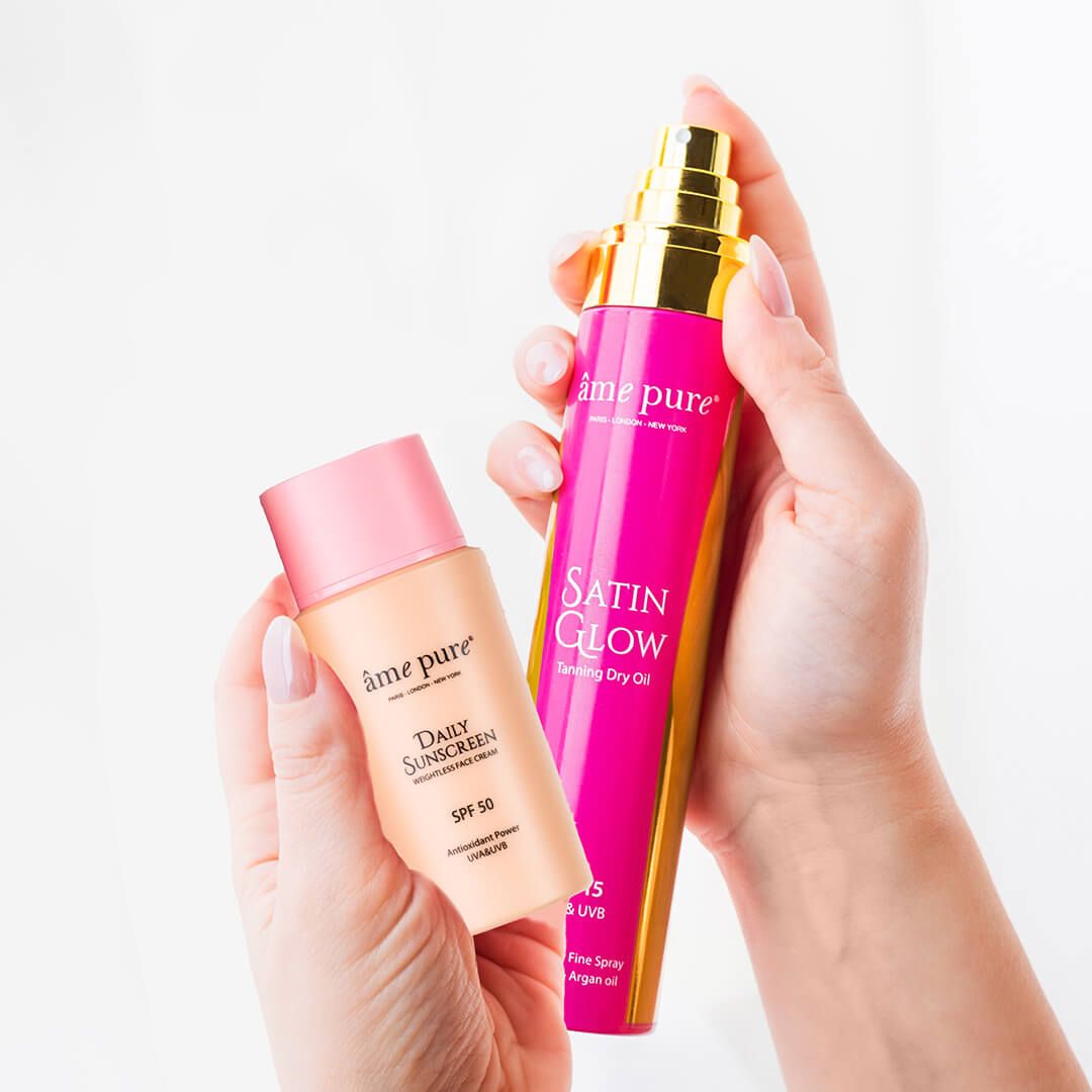 Mains tenant la crème solaire quotidienne SPF 50 et l'huile bronzante Satin Glow.