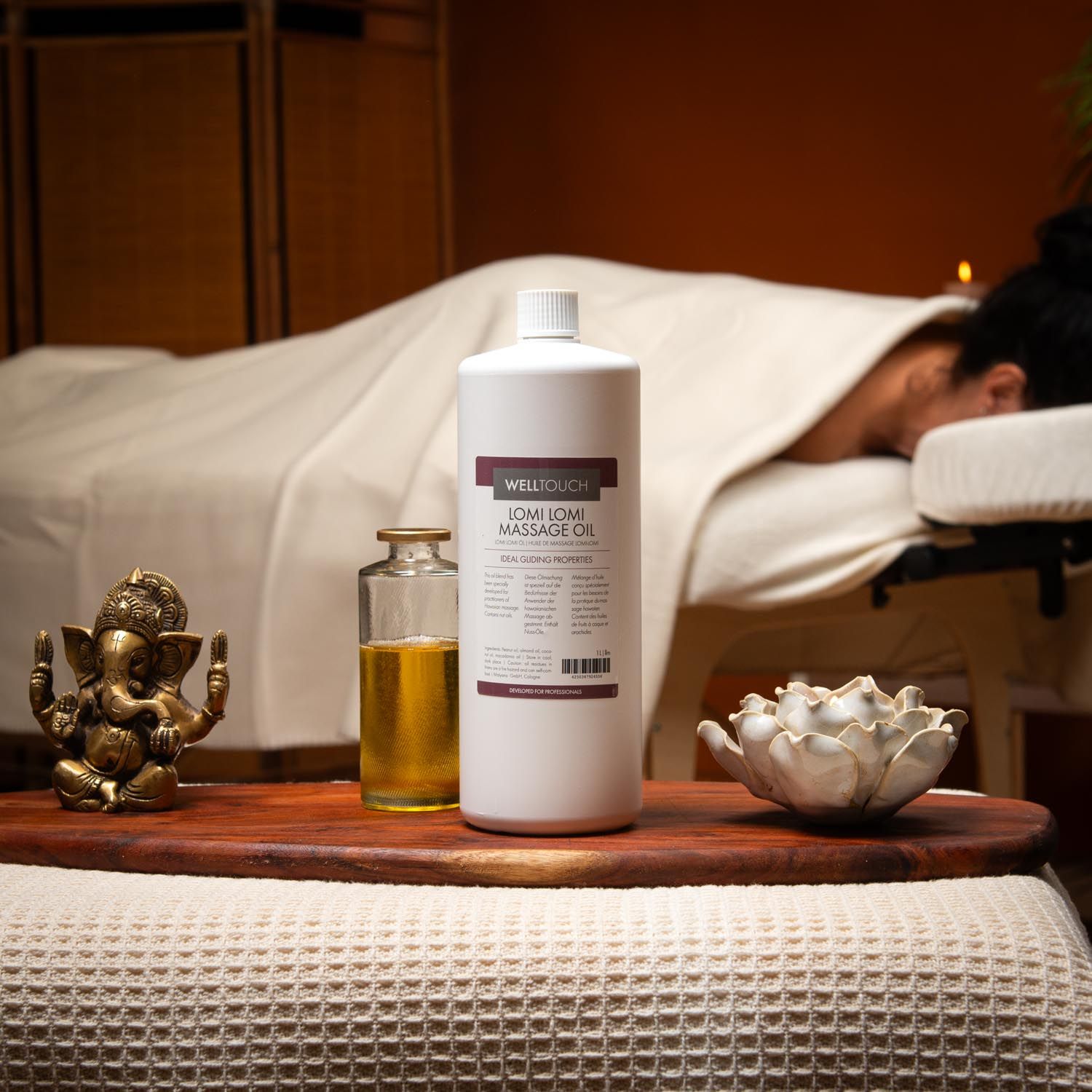 Grande bouteille blanche d'huile de massage Welltouch Lomi Lomi. À côté, une petite bouteille en verre avec de l'huile. En arrière-plan, une personne sur une table de massage.
