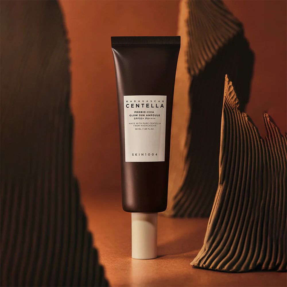 Tube marron avec étiquette blanche. Nom du produit et marque visibles. Placé sur un socle blanc, entouré d'éléments décoratifs.